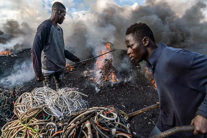 Een jonge man is afgebeeld die elektrische draden verbrandt om koper herwinnen in Agbogbloshie, Ghana, op het moment dat een andere metaalafvalverwerker arriveert met meer te verbranden draden. Een artikel uit 2018 in het "International Journal of Cancer" rapporteert een correlatie tussen nabijheid van verbrandingssites van e-afval en kinderlymfoom. (Image by Muntaka Chasant, gepubliceerd onder een CC-BY-SA-4.0 licentie.)